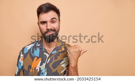 Serious attentive bearded man points thumb away recommends something shows copy space for your advertisement wears colorful shirt isolated over beige background. Special offer for you. Place for ad