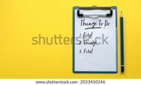 Top view of a pencil and a clipboard with paper written with Things To Do and a number with sentence First Things First on a yellow background with copy space. Royalty-Free Stock Photo #2033450246