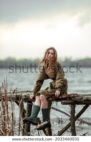 Lonely pensive girl, lost in thought, on a wooden bridge by the river, in a brown dress and rubber boots
