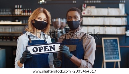 Portrait of cheerful mixed-race waiters standing in aprons and masks looking at camera in cafeteria and holding Open sign in hands. Working during coronavirus in coffee shop. Reopening concept