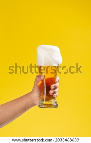 man's arm holds a glass beaker with beer inside, white foam and a solid yellow background, some foam falls Royalty-Free Stock Photo #2033468639