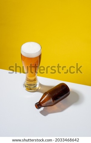cold beer served in a glass with white foam, an empty glass bottle on one side, on a white table and yellow background, no people Royalty-Free Stock Photo #2033468642