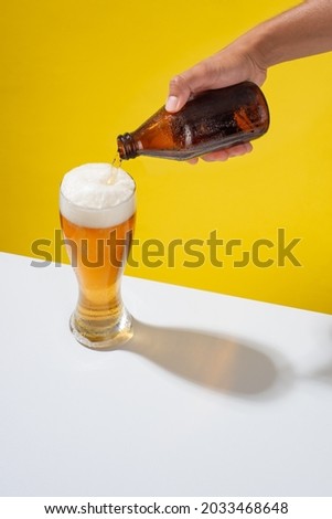 An arm serves a beer in a glass, with white foam, on a white table and yellow background Royalty-Free Stock Photo #2033468648