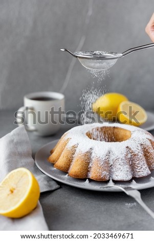 Sprinkling icing sugar over fresh homemade lemon pound cake. Powdered sugar in sieve. Cup of tea or coffee on the table. Home baked loaf cake. Classic recipe. Rustic. Simple grey background
