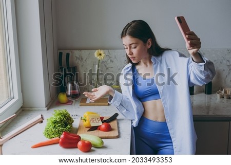 Young uncertain housewife woman 20s in casual clothes blue shirt talk by video call mobile cell phone show vegetable ingredients cook food in light kitchen at home alone Healthy diet lifestyle concept