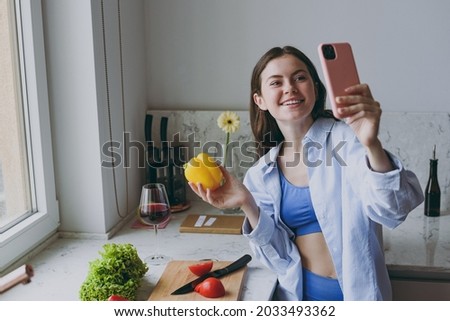Young housewife woman 20s in casual clothes blue shirt do selfie shot on mobile phone post photo social network hold bell pepper cook food in light kitchen at home alone Healthy diet lifestyle concept