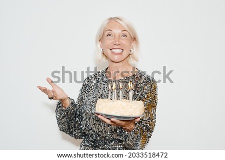 Waist up portrait of glamorous mature woman holding birthday cake against white background at party, shot with flash
