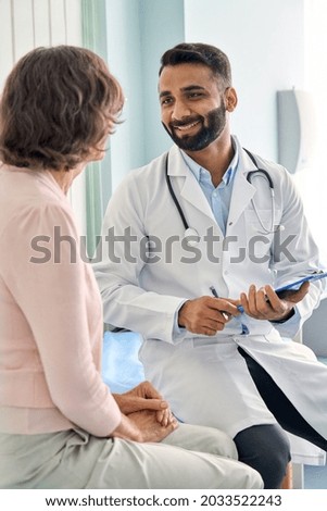 Vertical shot of young happy cheerful Indian medical worker therapist in white doctor's robe having appointment consulting older female patient in modern clinic hospital. Medical healthcare concept. Royalty-Free Stock Photo #2033522243