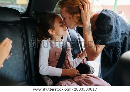 Mother giving her eight year old daughter a kiss. She's sitting on a back seat in a car. From inside a salon.