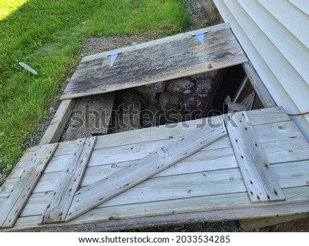 The side view of an open cellar door. The walls along the cement stairs are made of stone. There is also an empty hook that's meant to store a hose. Royalty-Free Stock Photo #2033534285