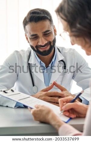Young happy Indian medical worker therapist in white robe having appointment consulting older senior patient sitting at desk filling data form in modern clinic hospital. Medical healthcare concept.