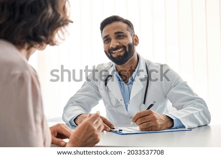 Young cheerful Indian doctor therapist in white robe having appointment with female patient sitting at table filling medical form in modern clinic hospital. Over shoulder view. Healthcare concept. Royalty-Free Stock Photo #2033537708