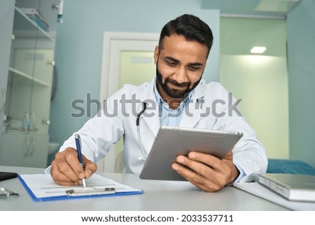Indian male happy general practice doctor therapist in modern clinic in white coat using tablet device having videocall, consulting remotely, writing health data. Telemedicine health care concept.