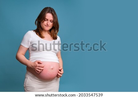 Portrait of a pregnant woman scratching her stomach, blue background Royalty-Free Stock Photo #2033547539