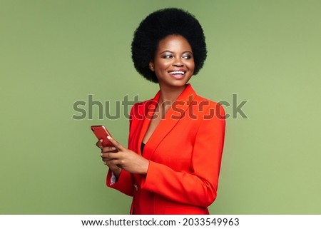 Successful african businesswoman hold smartphone look aside with confident happy smile. Afro american young entrepreneur female in luxury red suit with mobile phone isolated over green studio wall Royalty-Free Stock Photo #2033549963