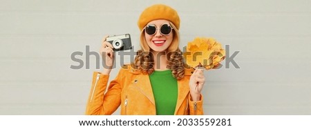 Autumn portrait of happy smiling young woman with film camera and yellow maple leaves wearing a french beret on gray background