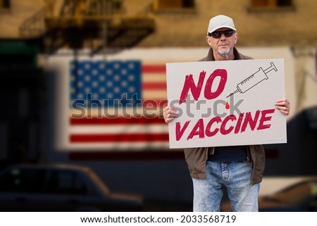 Displeased man with cap, blue jeans and sunglasses holding a NO COVID vaccine sign and a syringe with american stars and stripes flag on a wall in the background. Supporting anti-vaccination movement.