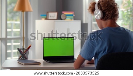 Back view of teenage student in headphones writing notes during web lesson on laptop at home. Green screen. Rear view of teen boy do homework or study remotely