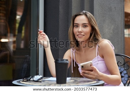 young asian pretty woman in pink dress sitting outdoors, writing notes in notebook, holding smartphone. attractive female student with cup of coffee outside of cafe studying. lifestyle, urban portrait