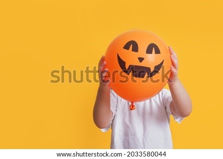 funny child in a white t-shirt holding an orange balloon with a picture of jack lantern, halloween mock up, yellow background copy space