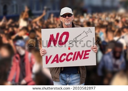 Displeased man with cap, blue jeans and sunglasses holding a NO COVID vaccine sign with crowd of people in background. Supporting anti-vaccination movement. Anti-vaxxer activist. 