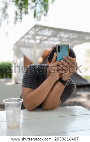 closeup man in causal t-shirt holding mobile phone up to taking photo during sit on white wooden table after drinking coffee in camping garden, relax lifestyle