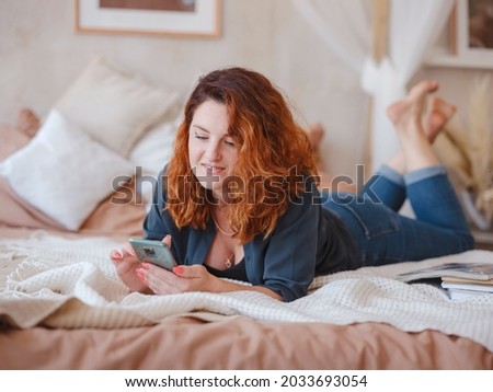 young redhead woman enjoys the morning silence in her room. Portrait beautiful lady using smartphone on bed in light interior.