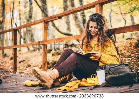  Brunette woman in yellow sweater sitting on a fallen autumn leaves in a park, reading a book or write a diary