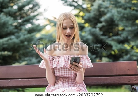 Attractive Caucasian young woman blonde shocked, surprised uses her mobile phone and read an interesting news sitting on a bench in the park in the warm summer.