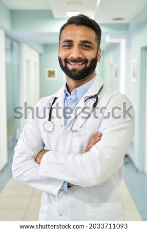 Smiling happy male Indian latin doctor medical worker wearing white coat with stethoscope around neck standing in modern private clinic with arms crossed looking at camera. Vertical portrait.