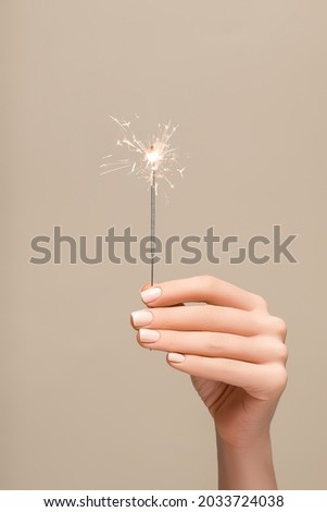 New Year lighted sparkler in female hand on beige background. Woman hold glowing holiday sparkling fireworks, shining fire flame. Christmas light. Royalty-Free Stock Photo #2033724038