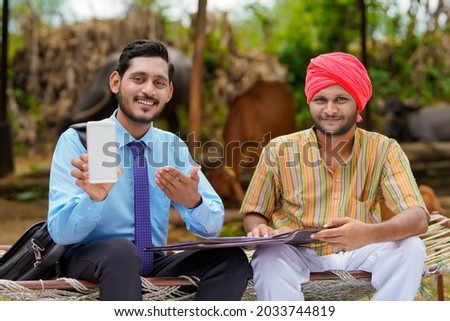 Young indian bank officer or agronomist showing smartphone with farmer at his farm