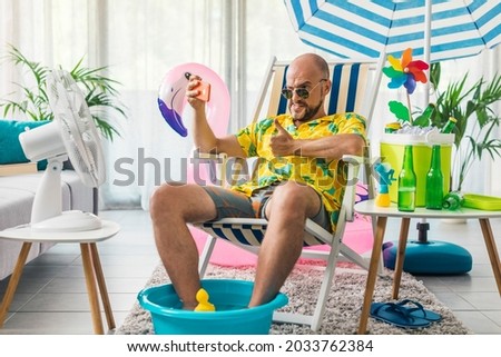 Happy man spending summer vacations at home and pretending he is on a beach, he is sitting on a deckchair and taking selfies with his smartphone