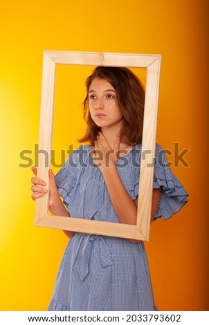 a cute teenage girl of 15 years old shows a smile and sadness through a wooden frame holding her in her hands on a yellow background. Knowledge Day