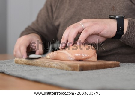 man with watch cutting a chicken breast on a board at the kitchen table
