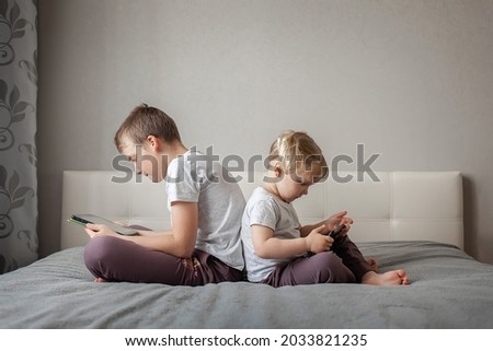 happy teenager little girl brother sister sitting on bed home with their backs each other and playing gadgets, tablet, phone, watching cartoons. concept modern childhood, home education, technology