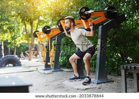 Open air gym. Street training on the municipal sports equipment. Athletic man doing squats using outdoor training machine. Royalty-Free Stock Photo #2033878844