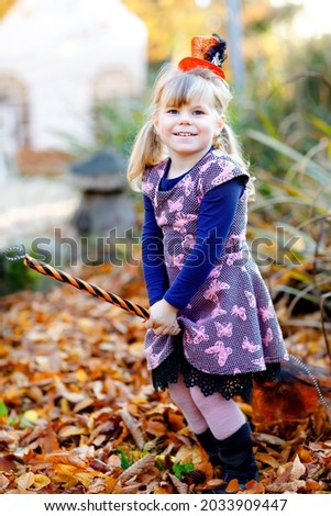 Cute little toddler girl dressed as a witch celebrates Halloween. Happy child outdoors, with orange funny hat and hold witch broom. Beautiful family festival season in october. Outdoor activity