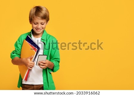 Cheerful european young blond boy pupil with backpack hold books and typing on smartphone, isolated on yellow background. Modern devices for study, education and knowledge, social distance and chat
