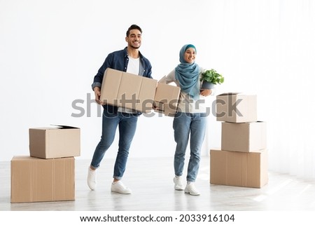 Excited arabic couple moving to new apartment, standing together over white wall with big paper boxes full of their belongings, copy space, full length photo. Moving, relocation, real estate concept