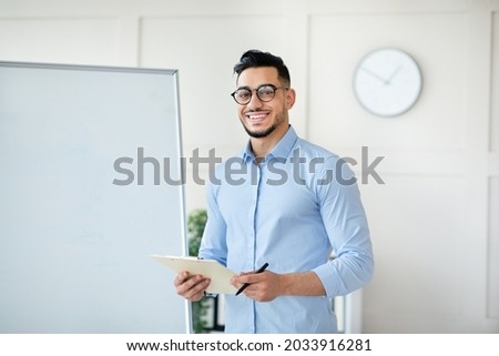 Online schooling. Positive Arab male teacher standing near empty blackboard with clipboard, giving remote lecture on web, speaking to students, mockup. Happy young tutor conducting lesson on internet Royalty-Free Stock Photo #2033916281