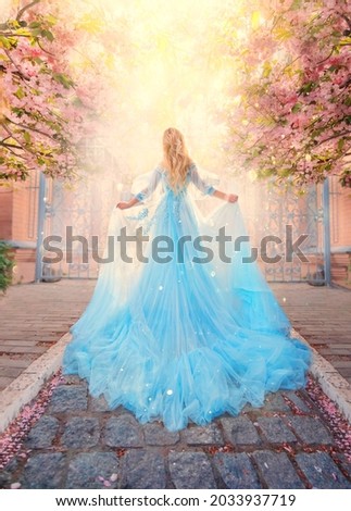 Art photo fantasy woman princess goes to meet light sun along spring street. Pink flowers sakura tree outdoor. Blue lush fairy dress, long train. Silhouette girl goddess, back view. Queen blond hair.