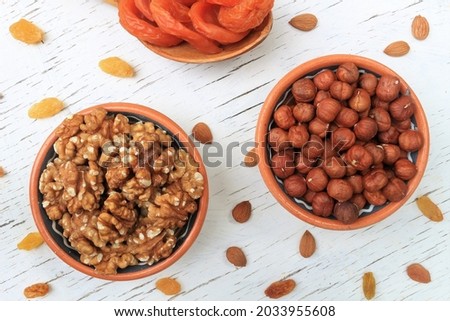 An illustration of healthy nutrition, organic food for those who take care of their health. Brown walnuts and huzelnuts in bowls, orange dried apricots in a spoon and yellow raisins on the white desk
