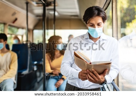 Portrait Of Handsome Millennial Guy In White Shirt And Face Mask Reading Book Standing In Bus And Leaning On Window. Focused Man Enjoying Longread Fiction Bestseller Novel. Read More Concept Royalty-Free Stock Photo #2033958461