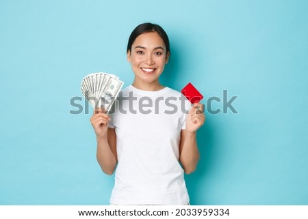 Shopping, money and finance concept. Smiling young pretty asian girl in white t-shirt, prefer to use both cash and credit card, showing you how make investment, standing light-blue background