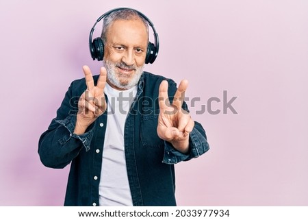 Handsome senior man with beard listening to music using headphones smiling looking to the camera showing fingers doing victory sign. number two. 