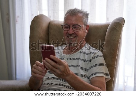 Older man is sitting in an armchair using his red smartphone with earphones. He wears glasses and a beard and a striped T-shirt. Behind him there are transparent curtains.