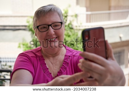 Older woman in pink t-shirt on her terrace at home. She is holding a red smartphone and taking a selfie.