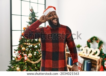 African american man wearing santa claus hat standing by christmas tree doing peace symbol with fingers over face, smiling cheerful showing victory 