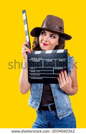 Studio portrait of a young attractive woman with a brown hat and a jeans outfit holding a movie clapperboard against a bright vivid yellow background. Breezy and dreaming look.
